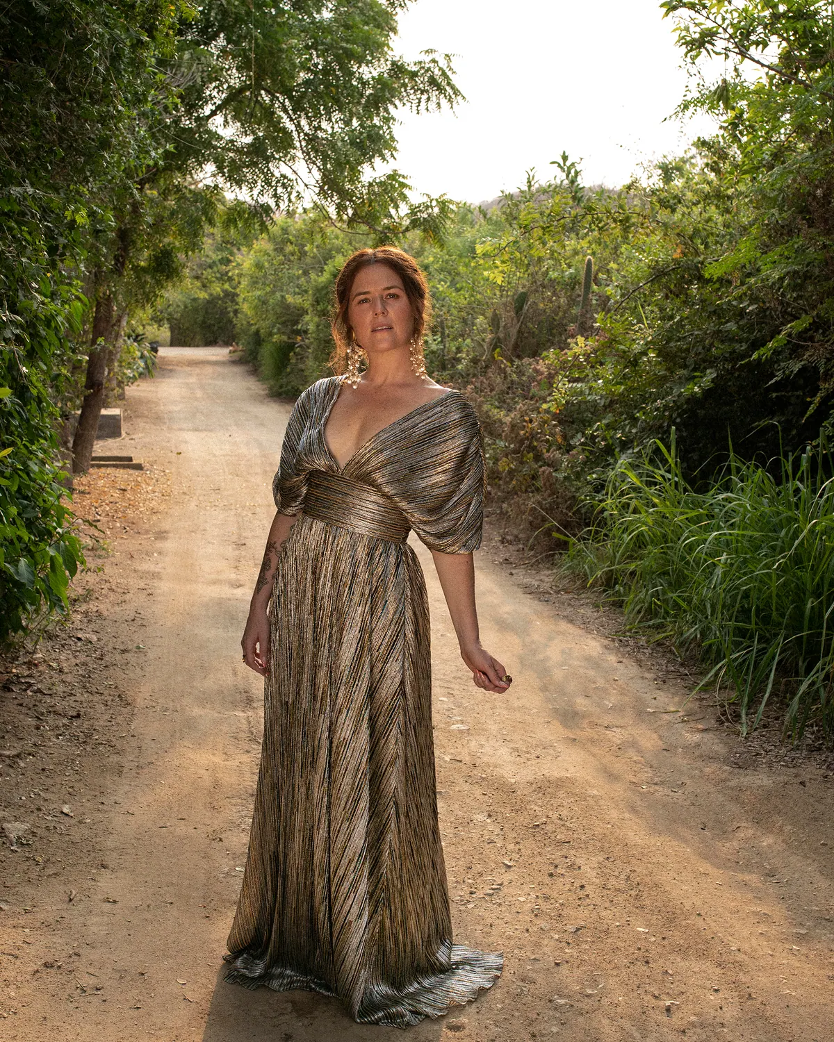 iskwē standing on a dirt path between green trees in a metallic gold gown