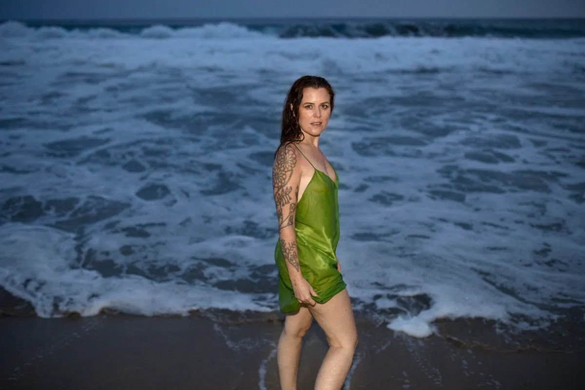 iskwē standing in the surf at dusk in a green slip dress, looking back over her shoulder
