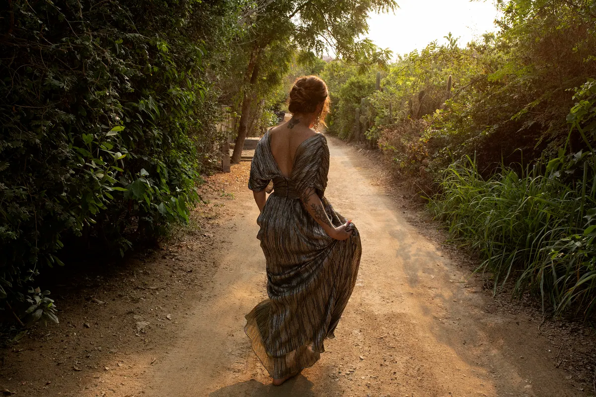 iskwē walking away down a sunlit dirt path in a flowing gown